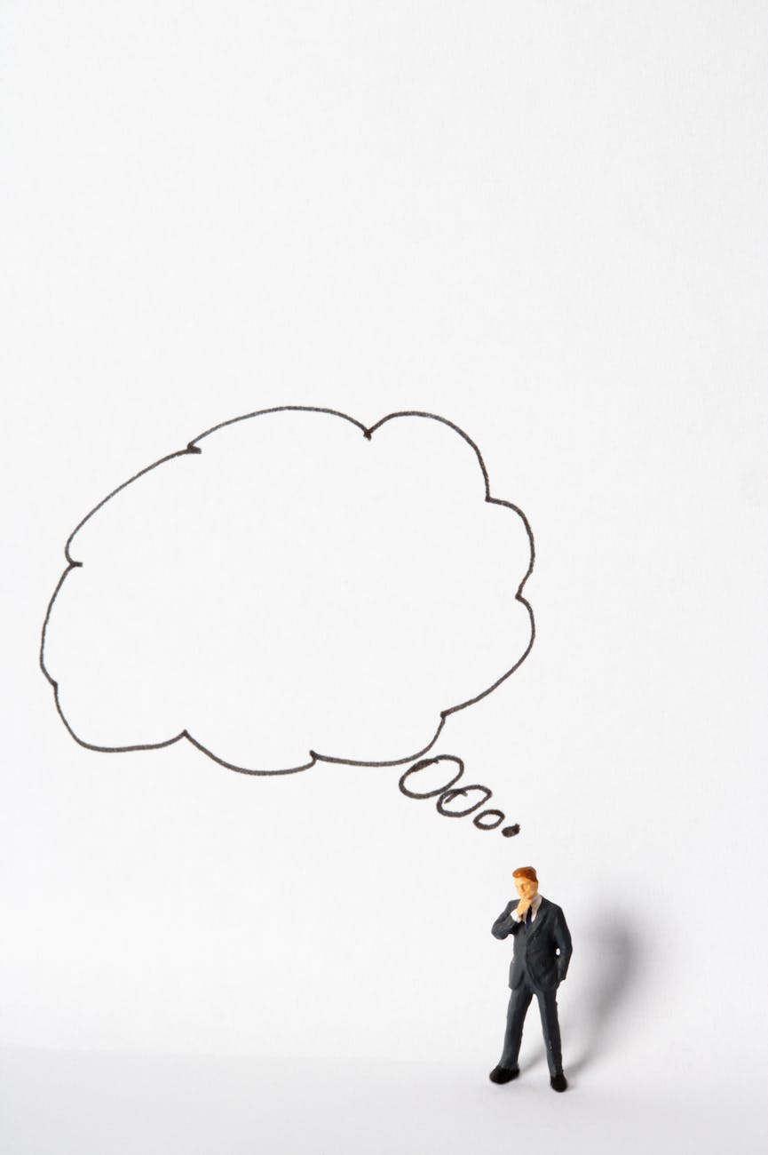 A miniature businessman stands pondering under a large thought bubble on a white background.