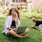 Woman multitasking on phone and laptop while child plays outdoors.