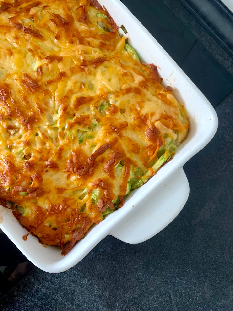 Delicious homemade cheesy baked casserole with vegetables in a white dish on dark countertop.