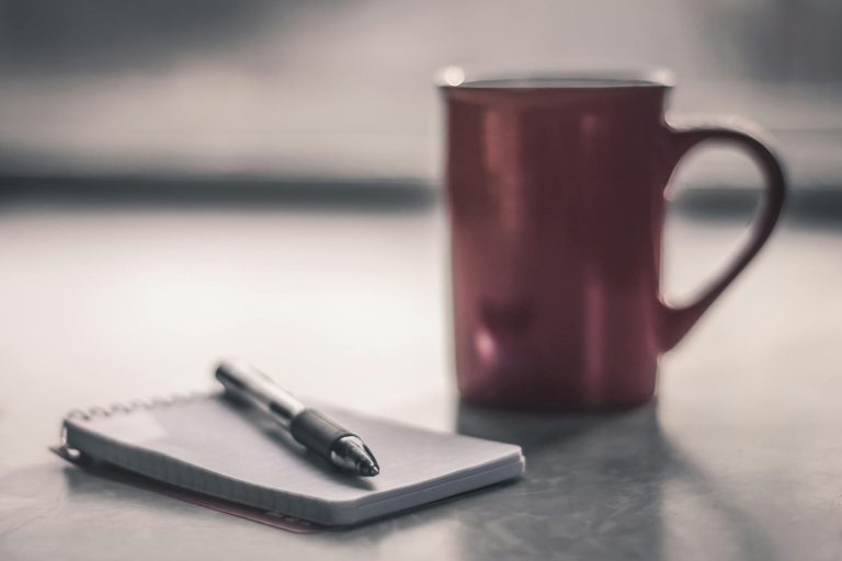 Blurred background image of a cup and a notepad with a pen on a desk, perfect for creative inspiration.