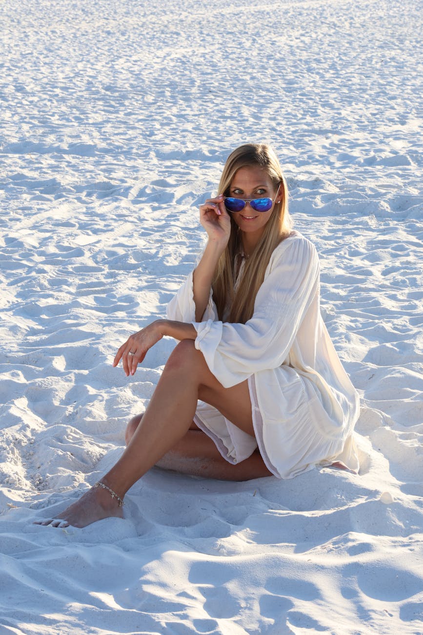 A woman in a white dress sits on white sand, wearing sunglasses, enjoying a sunny day outdoors.