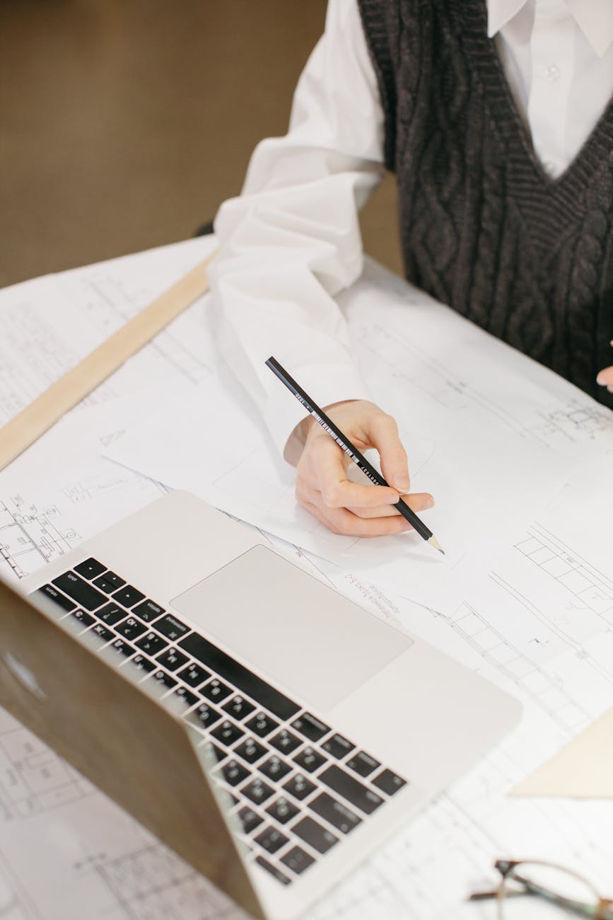 A close-up of an architect sketching plans with a pencil beside a laptop.
