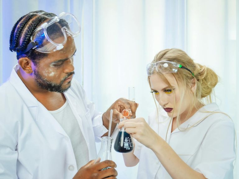 Two scientists in lab coats conducting a chemistry experiment using glasswares. Safety goggles worn for protection.