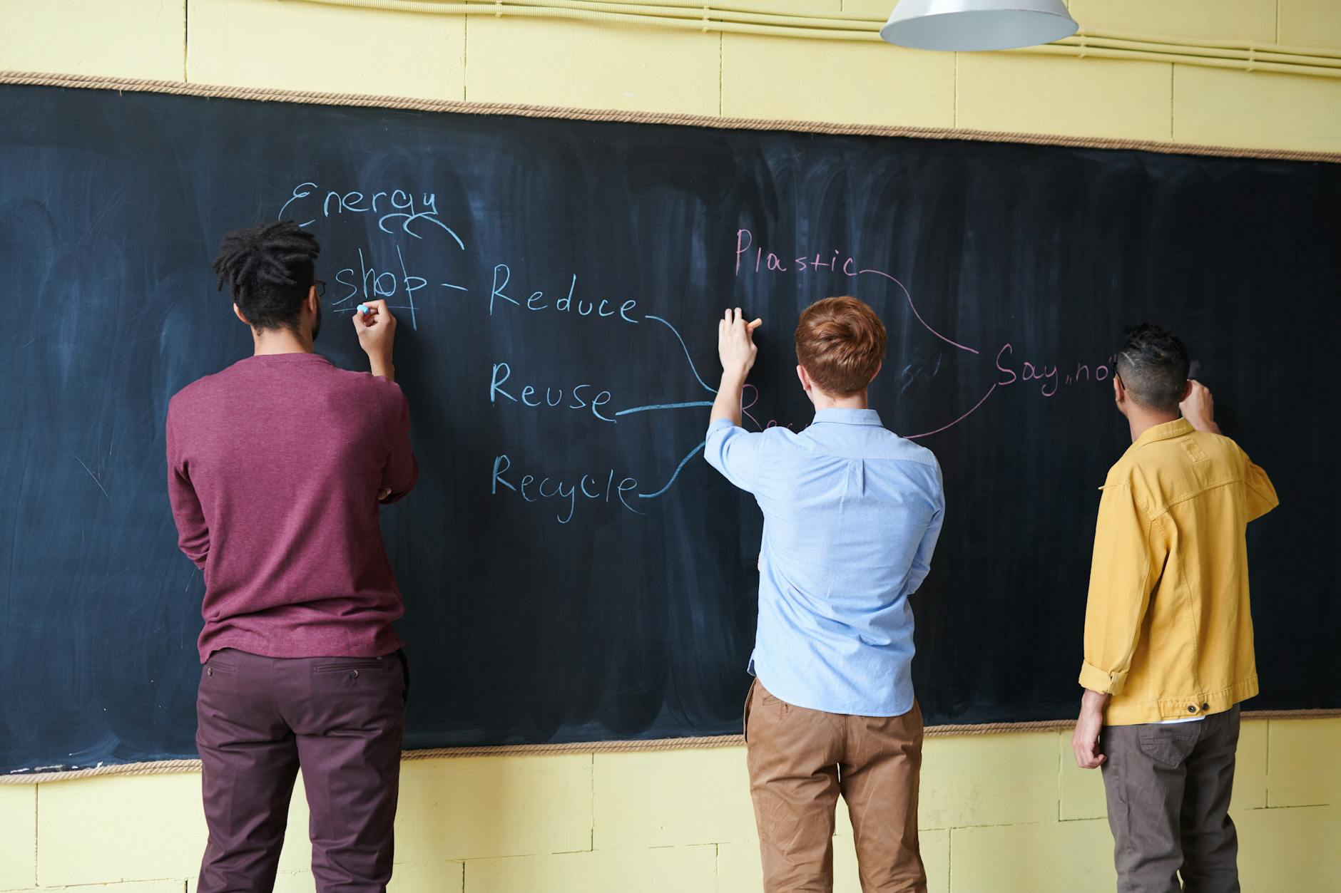 Three students writing on a chalkboard, focusing on reducing plastic and energy waste.