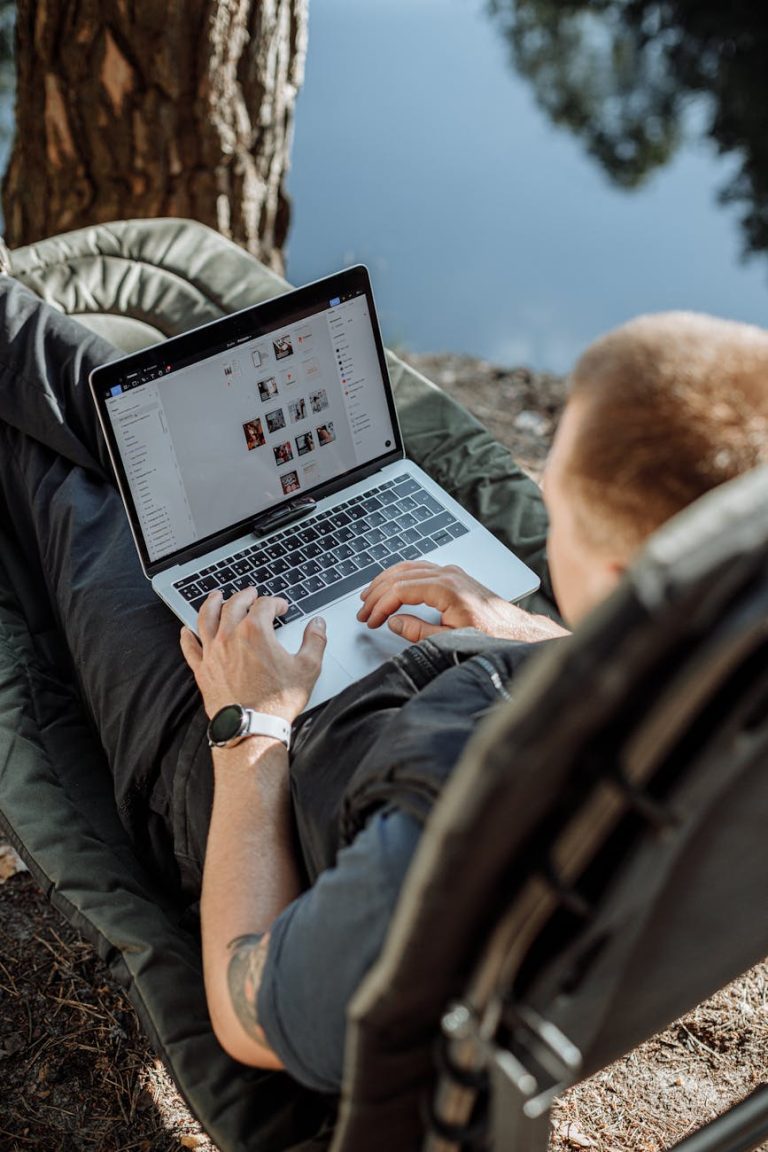 Young man working on a laptop outdoors by a lake, enjoying a serene camping setting.