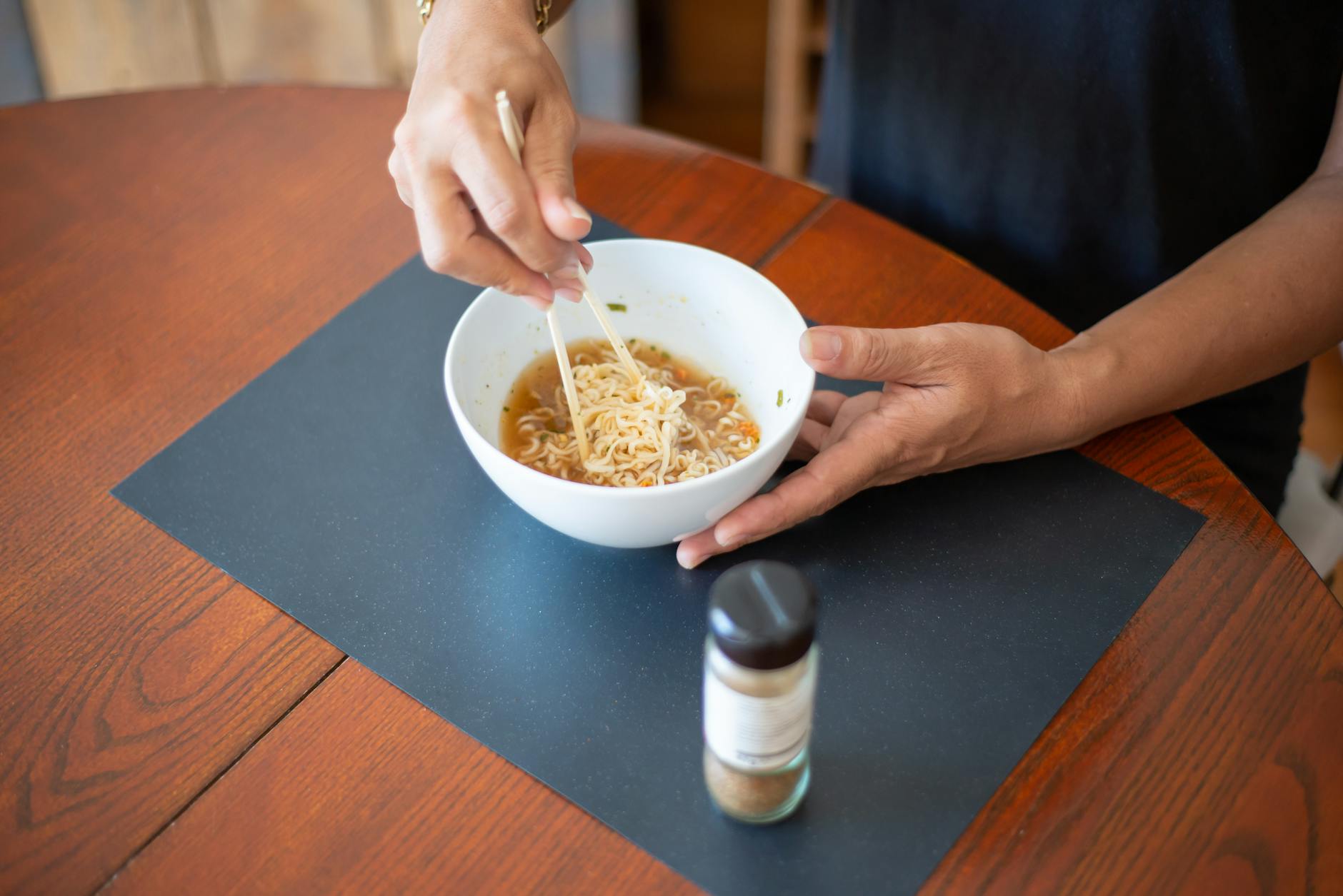 High-angle view of hands holding a bowl of Asian noodles with chopsticks on a wooden table.