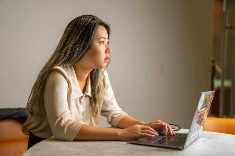 Young Asian woman concentrating on work, sitting at a desk using a laptop in an office setting.
