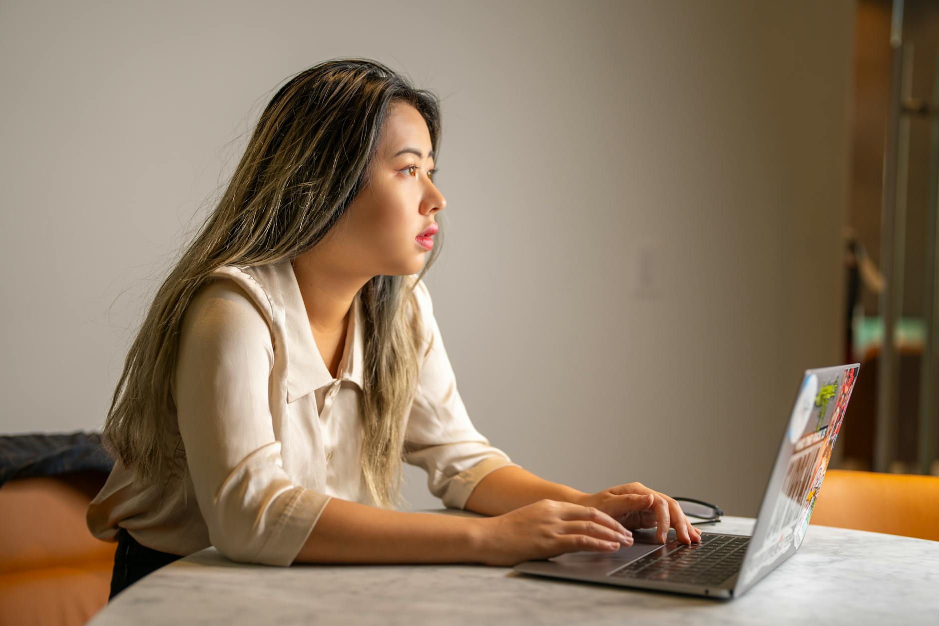 Young Asian woman concentrating on work, sitting at a desk using a laptop in an office setting.
