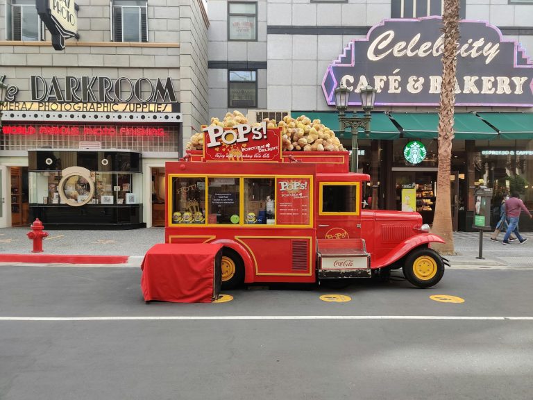 Colorful vintage popcorn truck outside cafes and shops on a bustling street.