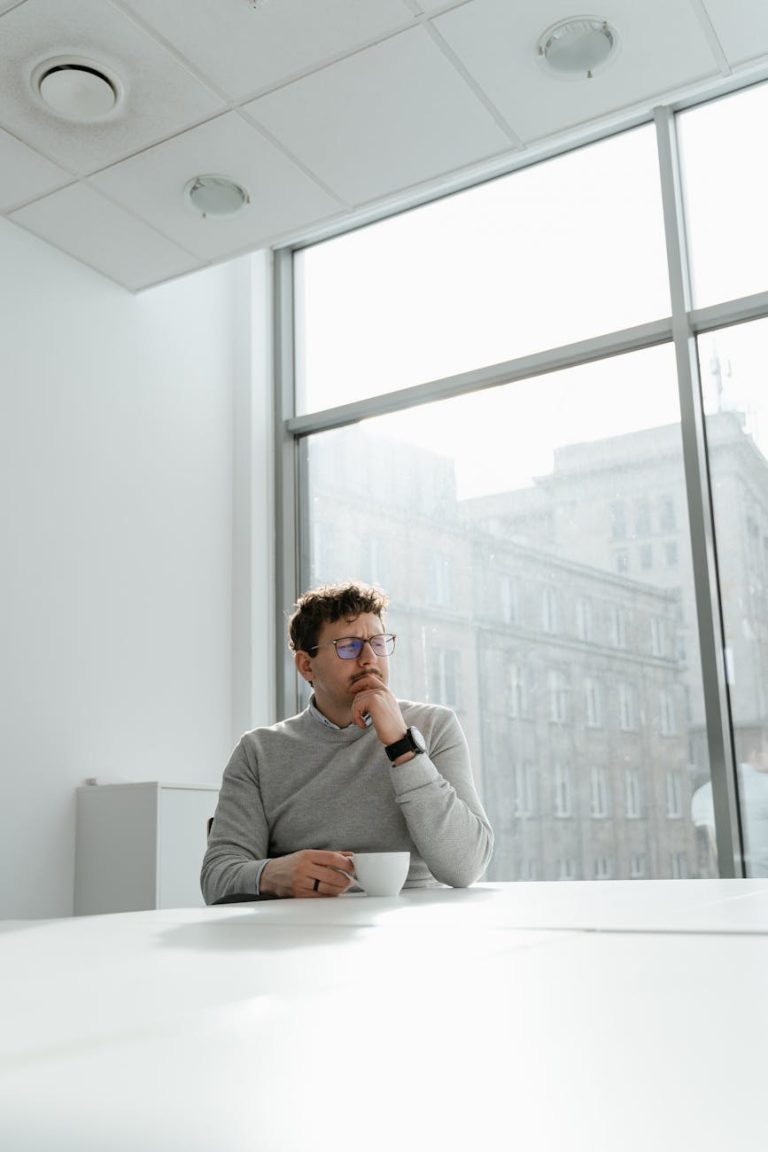 A thoughtful young businessman with glasses sits at a modern desk in a bright office.