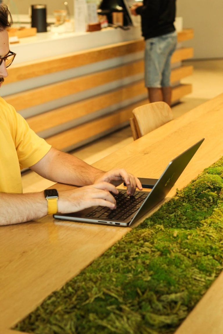 A person using a laptop at a wooden table with moss in a modern cafeteria setting.
