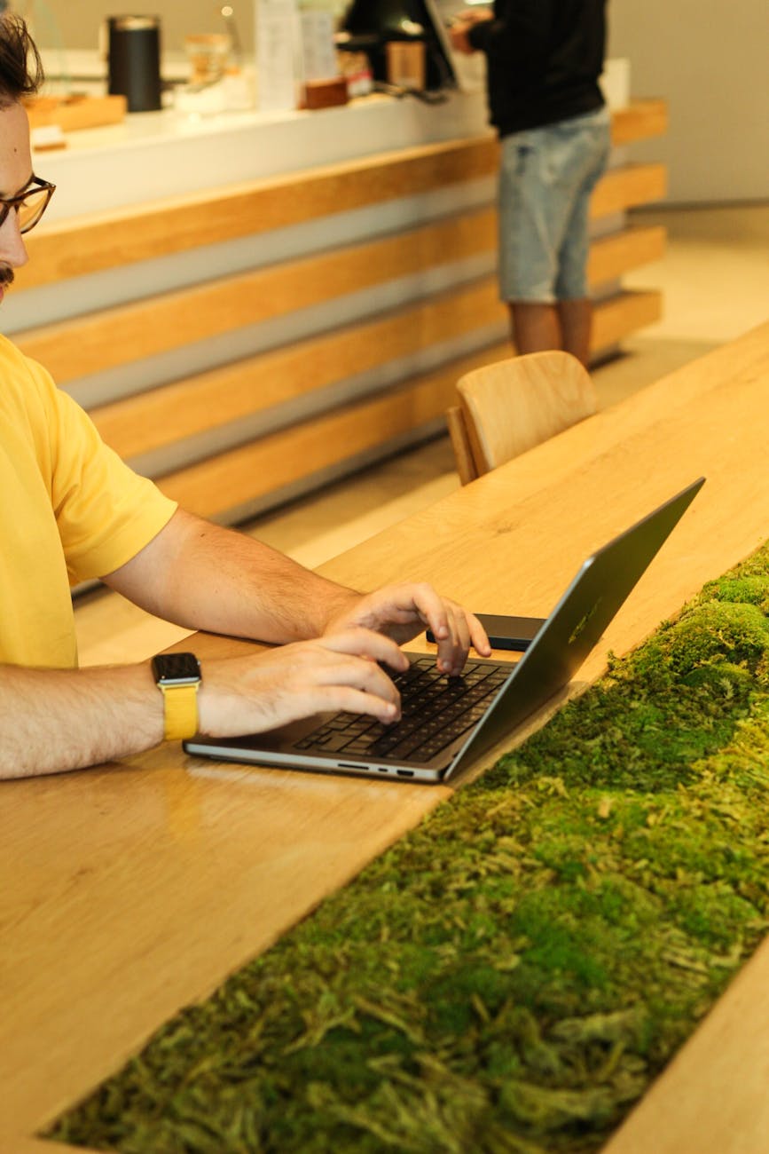 A person using a laptop at a wooden table with moss in a modern cafeteria setting.