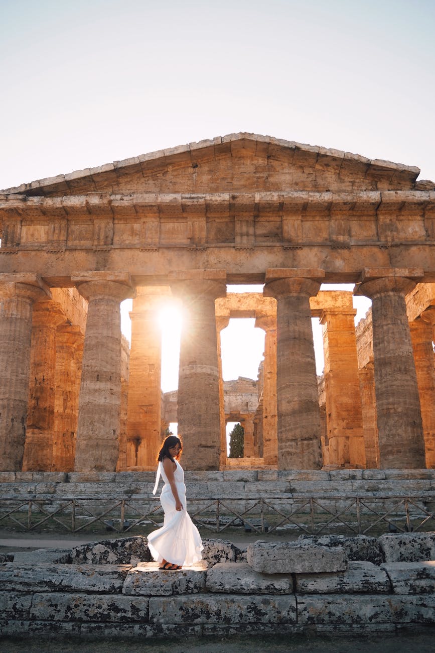 A woman in a white dress walks by an ancient temple at sunset, showcasing its grandeur.