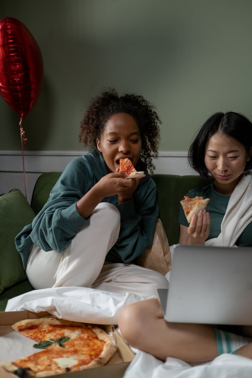 Two friends enjoying pizza and watching a movie together on a couch, creating a cozy atmosphere.