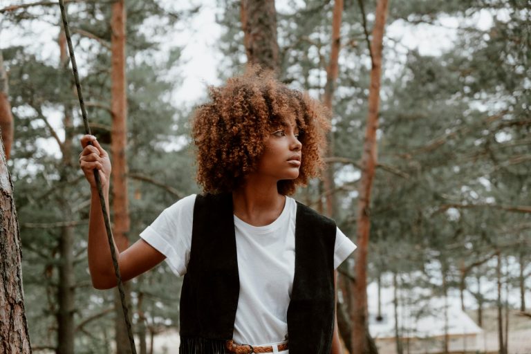 Stylish woman with curly hair stands in a forest, holding a rope and looking thoughtful.
