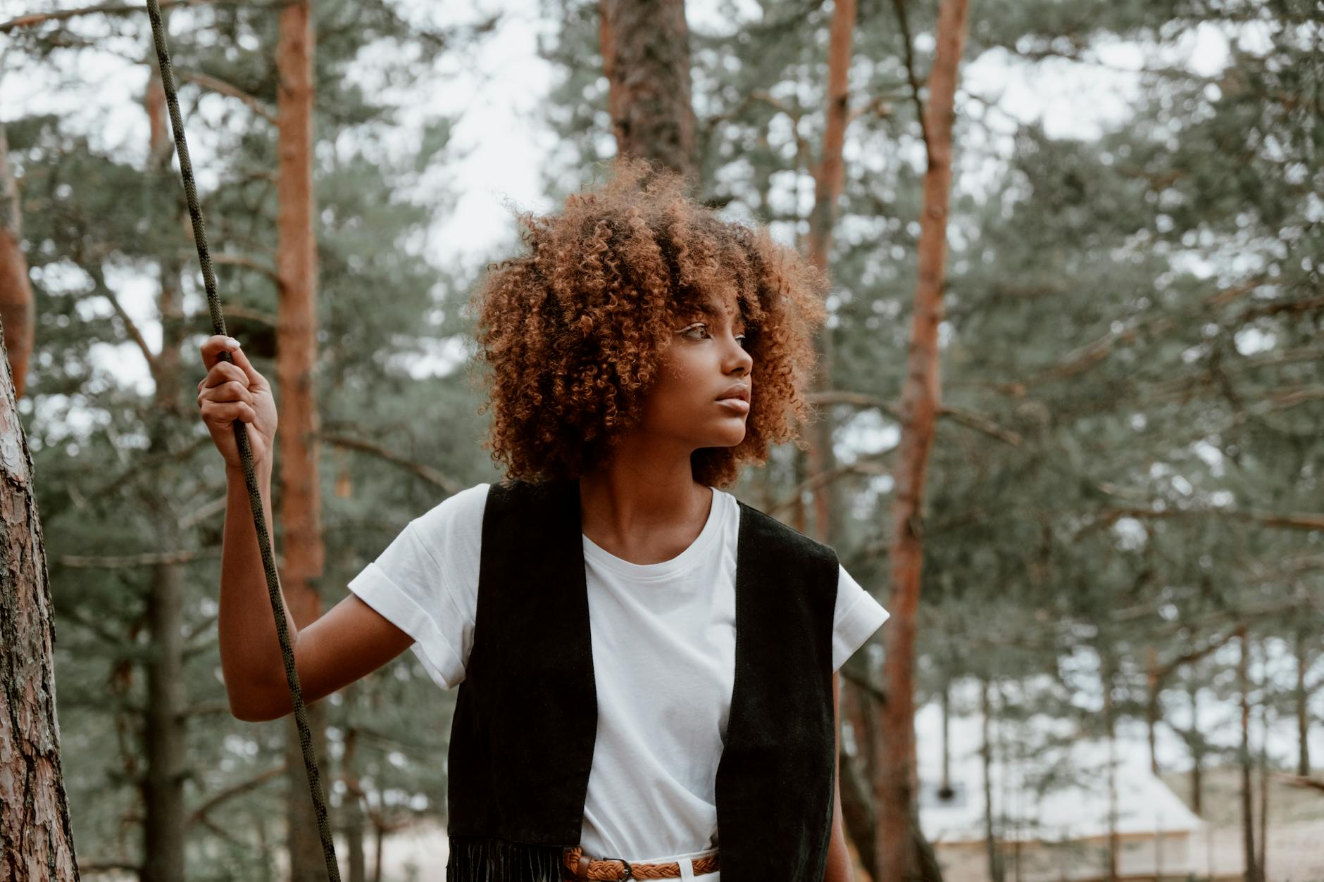 Stylish woman with curly hair stands in a forest, holding a rope and looking thoughtful.