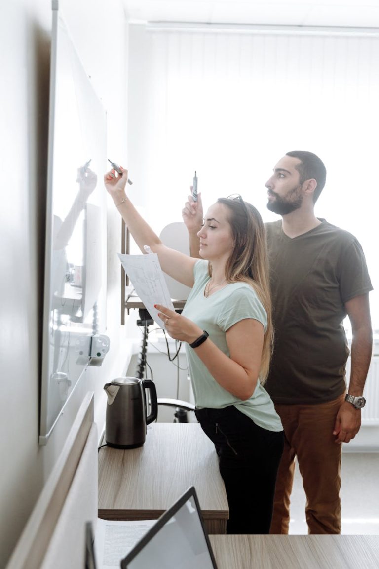 Two adults collaborate, writing on a whiteboard in a bright, modern office.