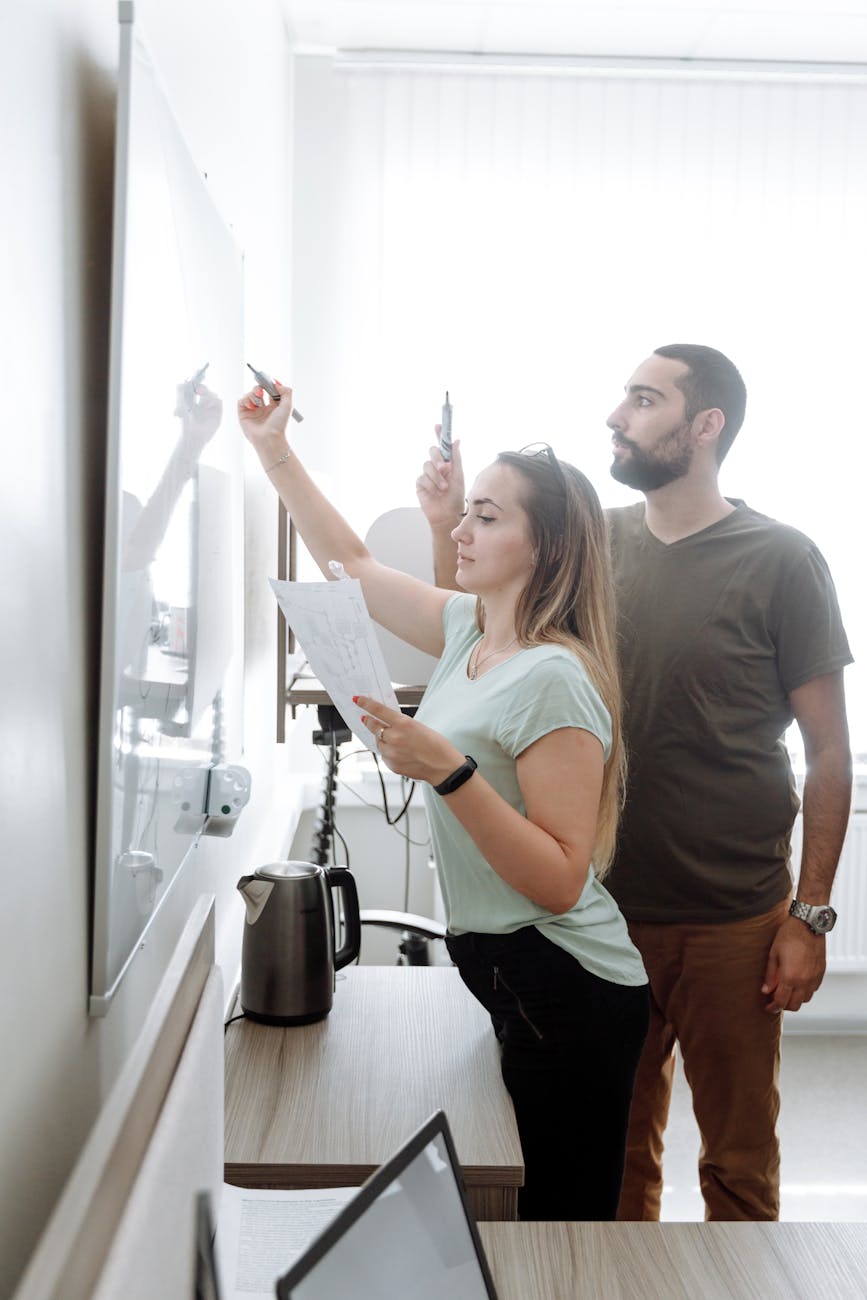 Two adults collaborate, writing on a whiteboard in a bright, modern office.