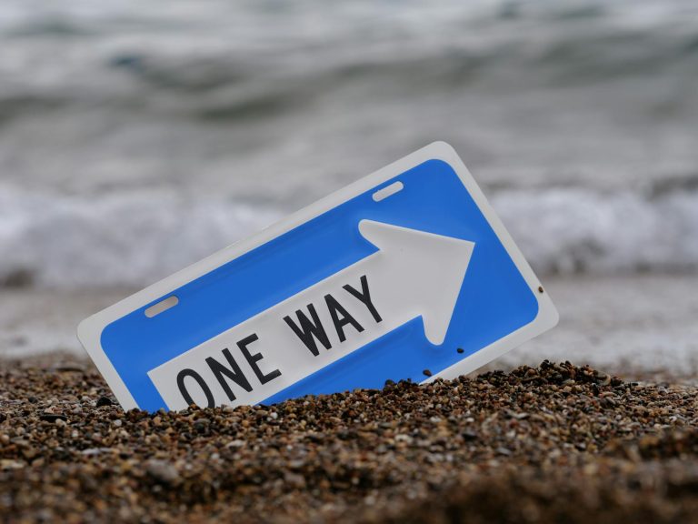 A blue one way sign in the sand by the ocean, with waves in the background.