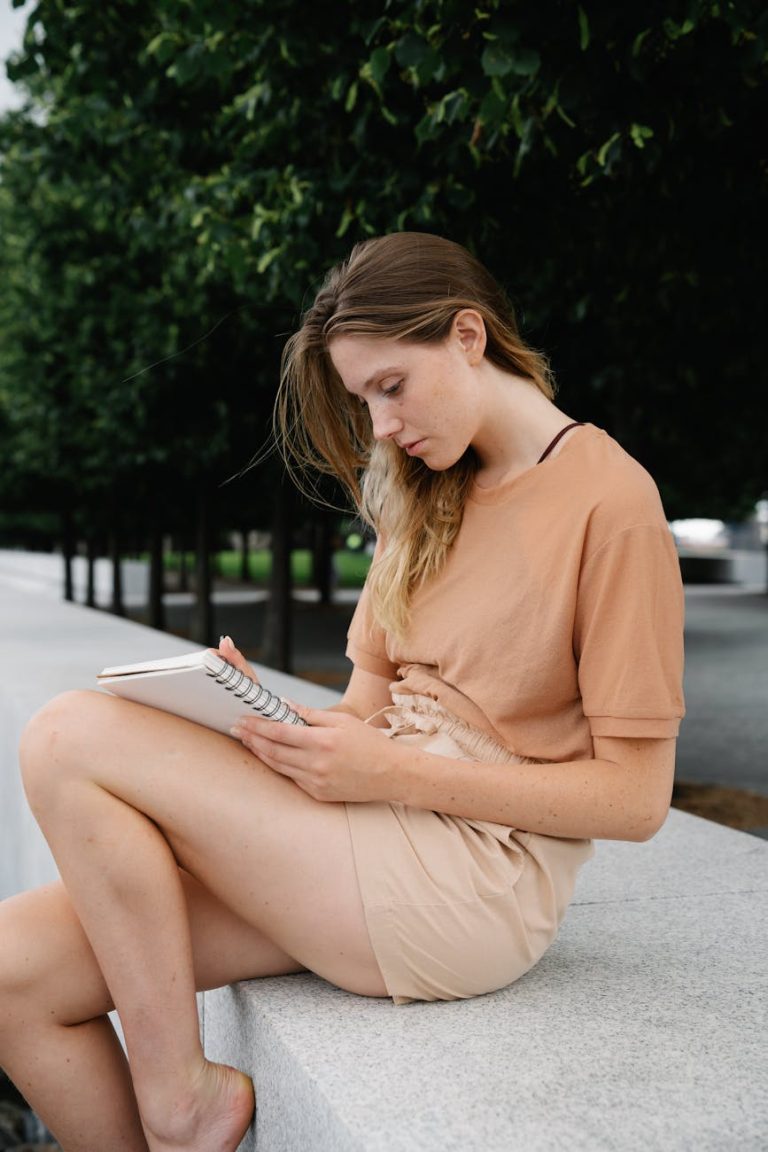 A young woman in a brown shirt sits outdoors on a concrete surface, writing in a notebook on a summer day.