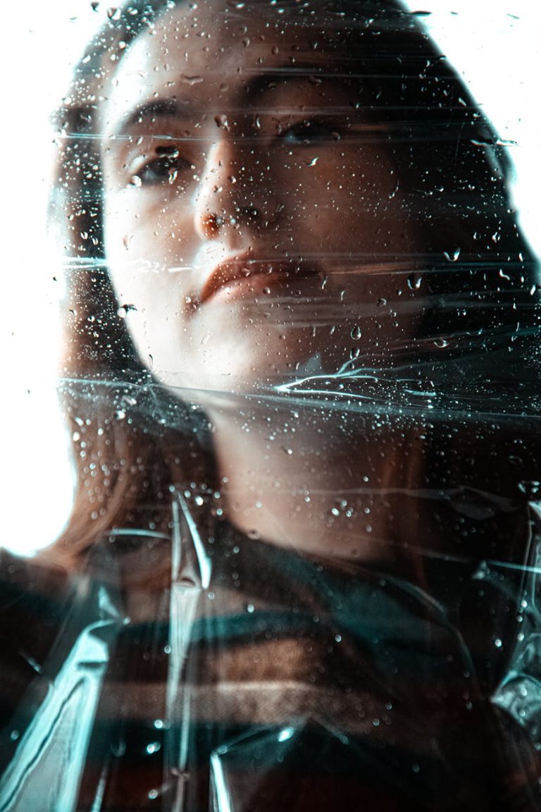 Intimate portrait of a young woman looking through a rain-speckled window, capturing a moody atmosphere.