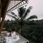 From above side view of anonymous female traveler in white blanket relaxing in hammock and admiring view of lush dense woods