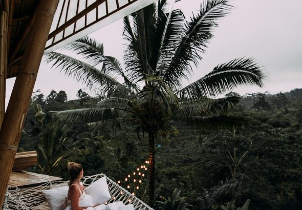 From above side view of anonymous female traveler in white blanket relaxing in hammock and admiring view of lush dense woods