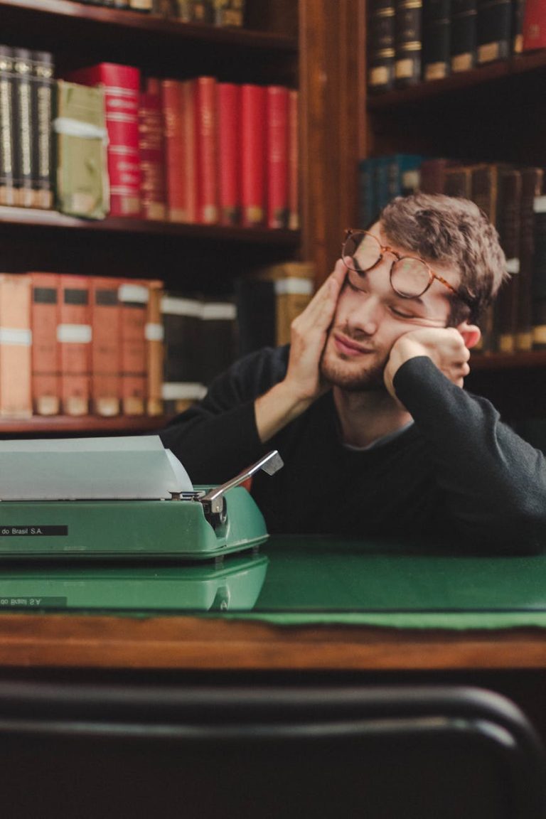 Tired man wearing glasses resting in a library with a typewriter, surrounded by old books.