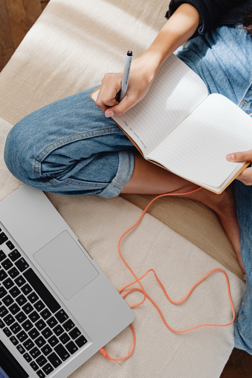A person in casual attire writing in a notebook beside a laptop, captured indoors.