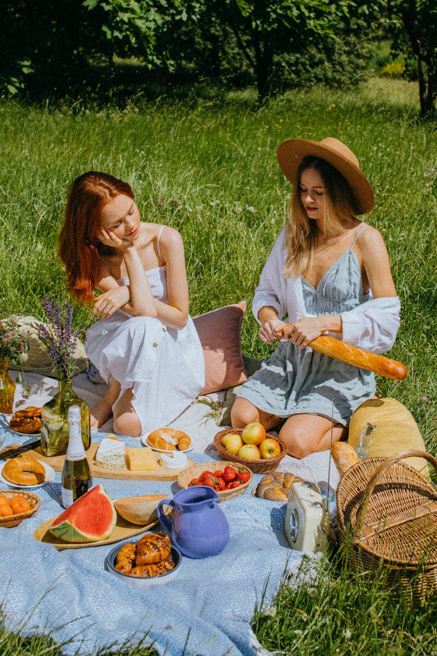 Two women enjoying a sunny picnic with fruits, bread, and wine in a lush park.