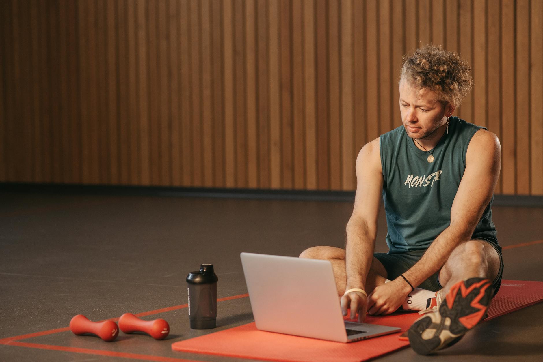 Man sitting on exercise mat with laptop, surrounded by dumbbells, creates a unique fitness workspace.