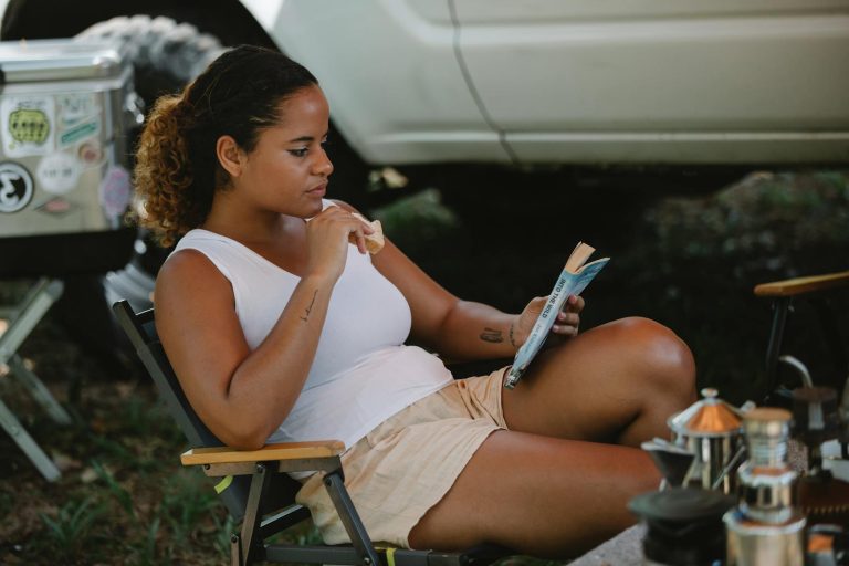 Young woman relaxing outdoors, reading a book and eating a snack during a summer camping trip.