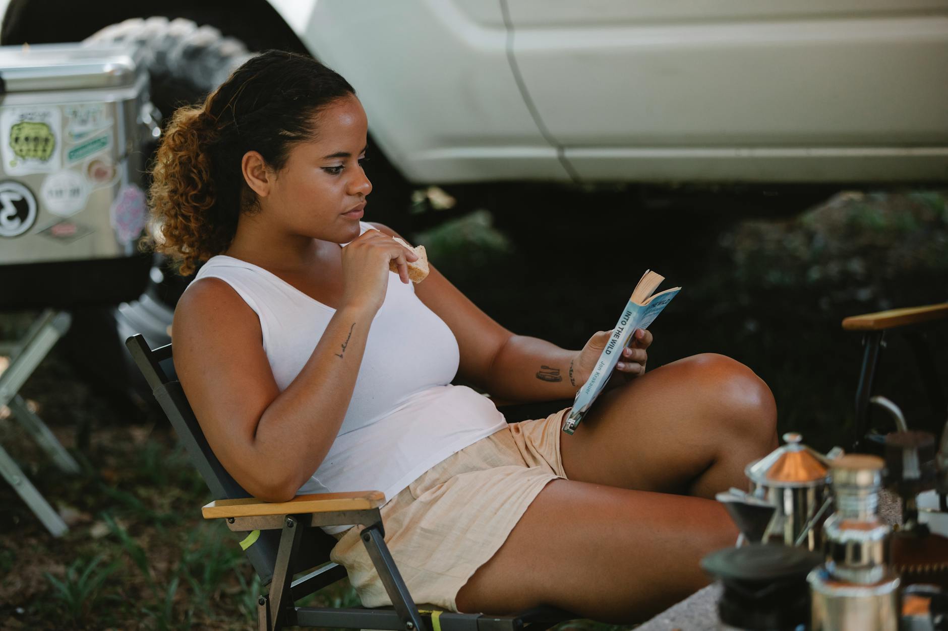 Young woman relaxing outdoors, reading a book and eating a snack during a summer camping trip.